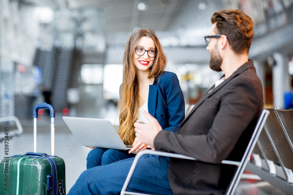 Elegant business couple sitting with laptop, phone and suitcase at the waiting hall in the airport. 