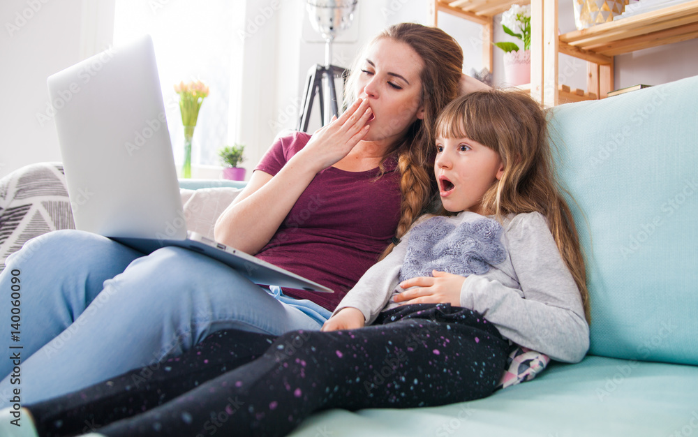 Mother yawning while excited daughter watching movie on laptop