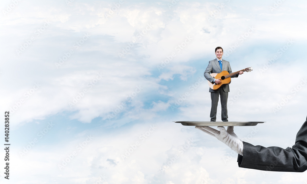 Businessman on metal tray playing acoustic guitar against blue sky background