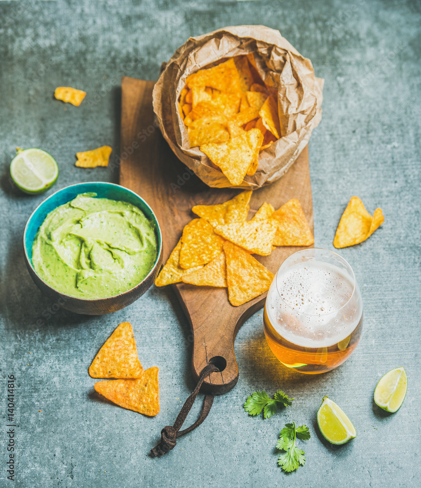 Mexican corn chips, fresh guacamole sauce and glass of beer on wooden serving board over grey concre