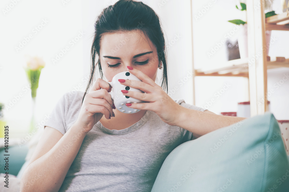 Young relaxed woman drinking coffee while sitting on comfortable sofa