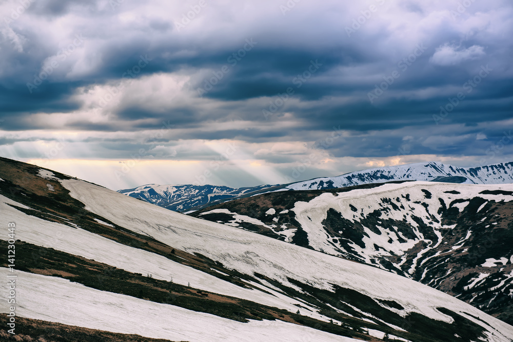 Spring mountain landscape with snow and grass. Dramatic clouds lying on the horizon and sun is shini