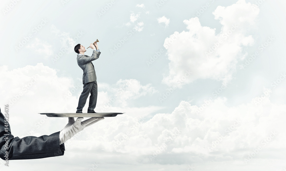 Businessman on metal tray playing fife against blue sky background