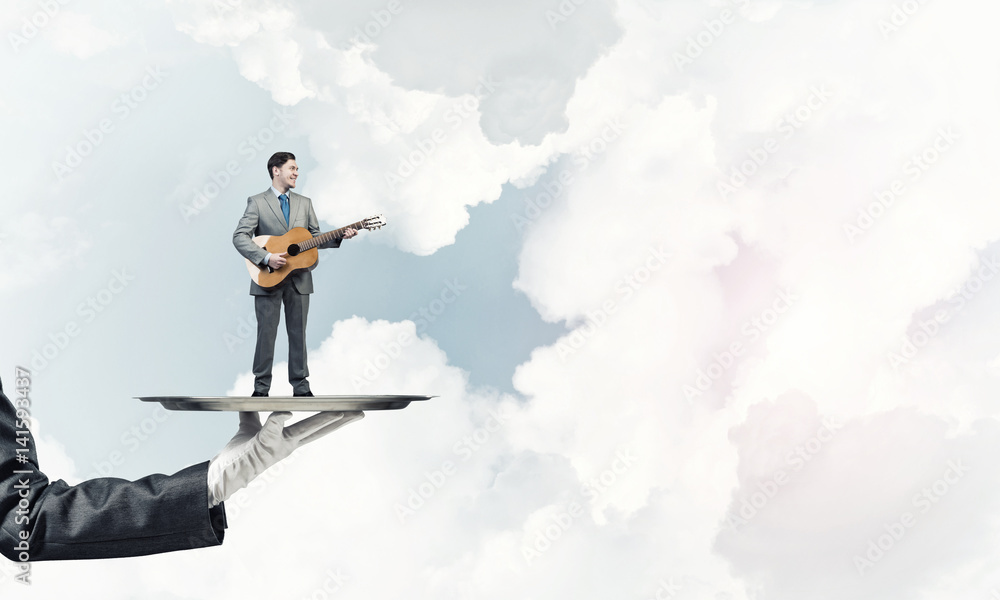 Businessman on metal tray playing acoustic guitar against blue sky background