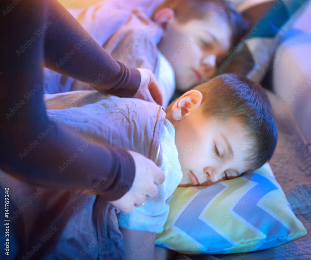 Children sleeping and dreaming in bed. Mother cover her little sons with a blanket