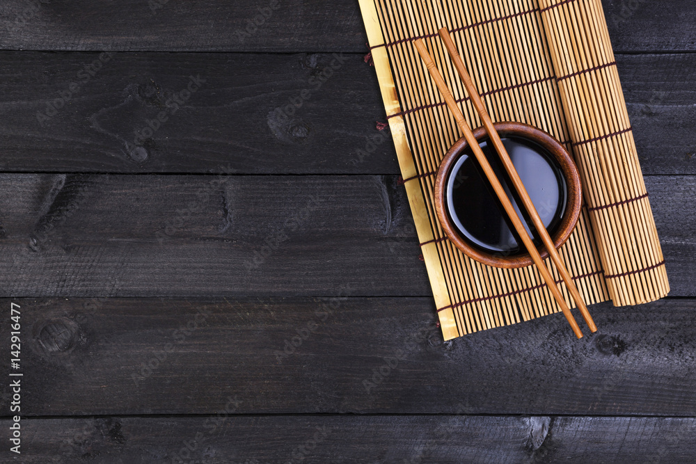 Background for sushi. Bamboo mat and soy sauce on black wooden table. Top view with copy space