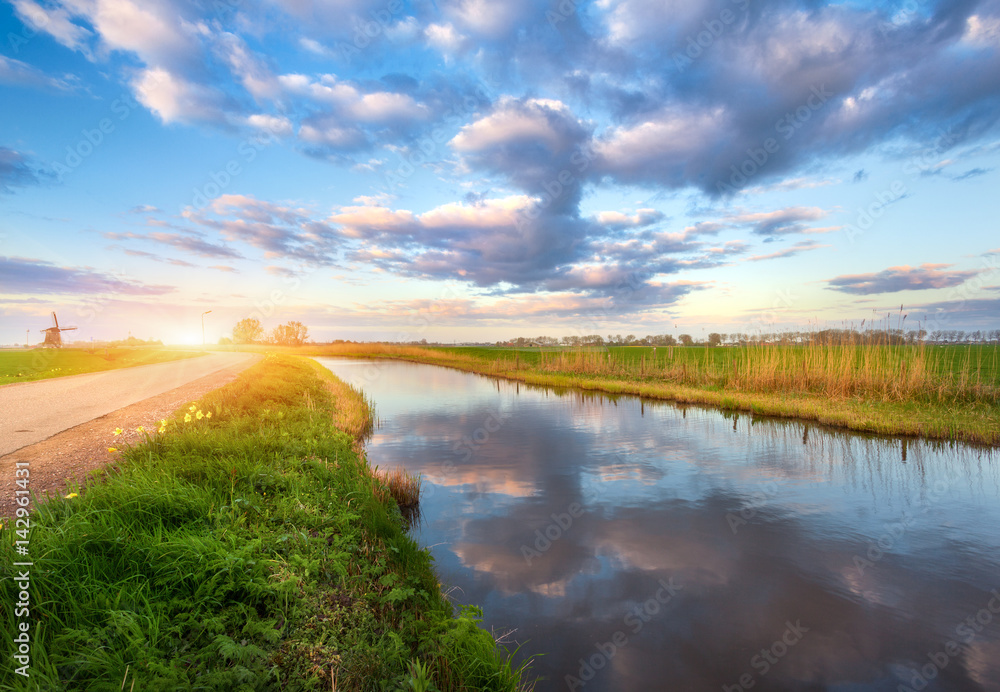 Spring landscape. Colorful blue cloudy sky reflected in water, green grass and yellow sun, road and 