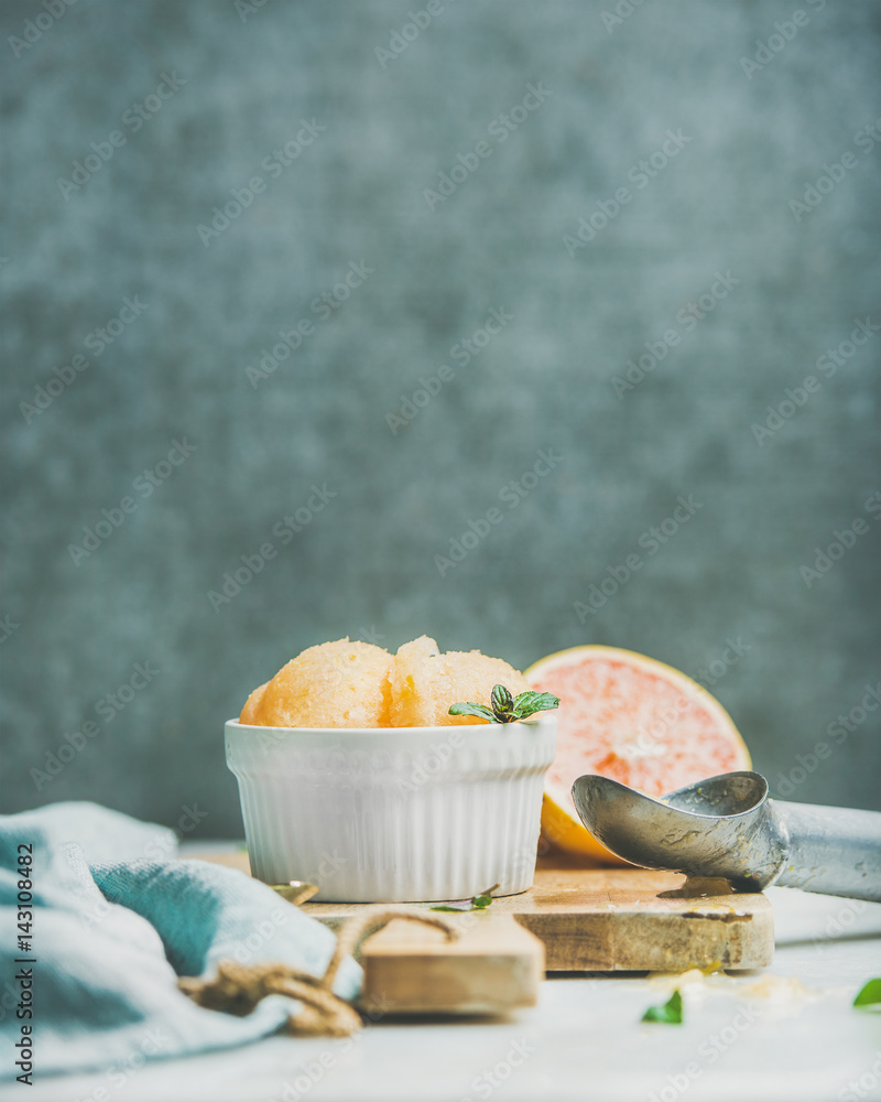 Pink grapefruit homemade sorbet with fresh mint leaves in white bowl on wooden board, grey plywood w