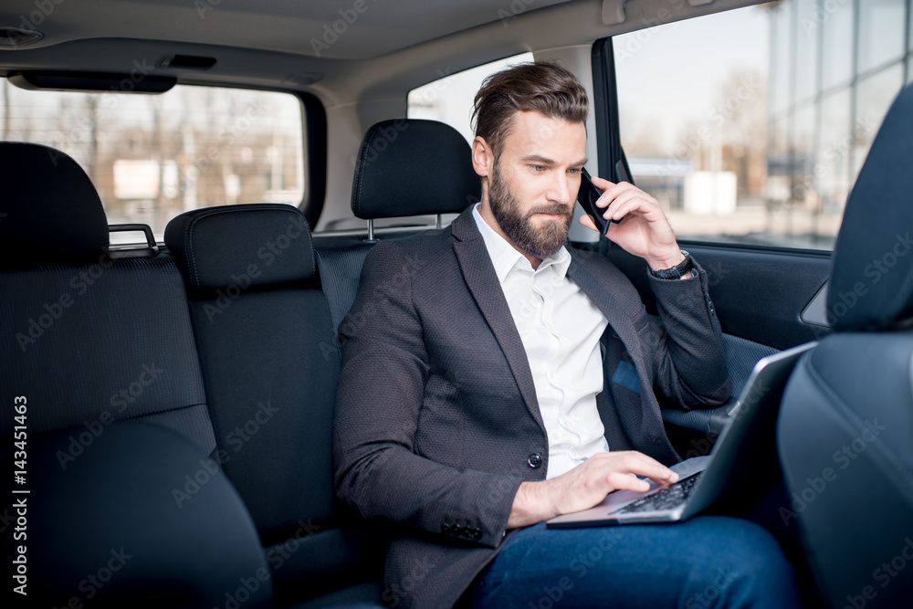 Handsome businessman talking with phone sitting with laptop on the backseat of the car