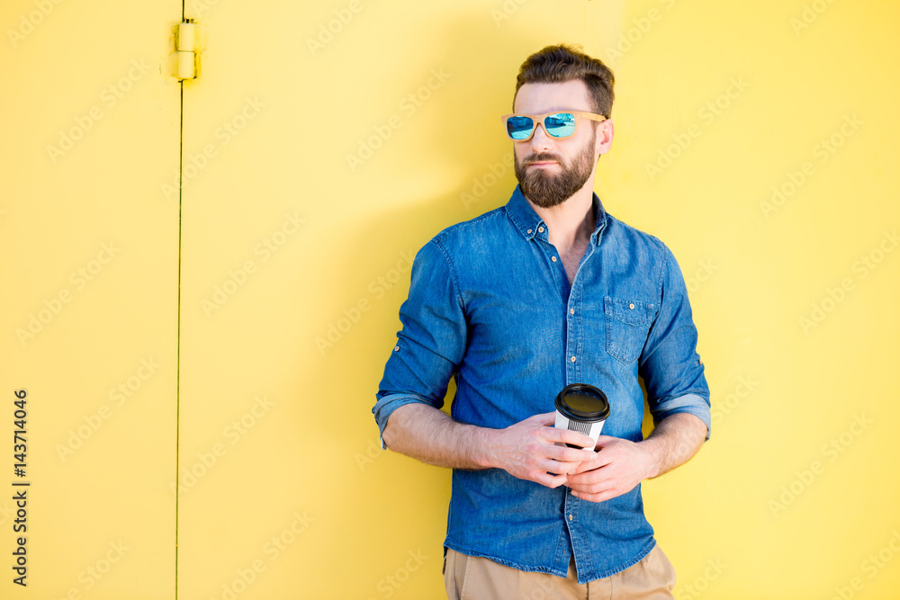 Portrait of a handsome man in blue t-shirt standing with coffee to go on the yellow background