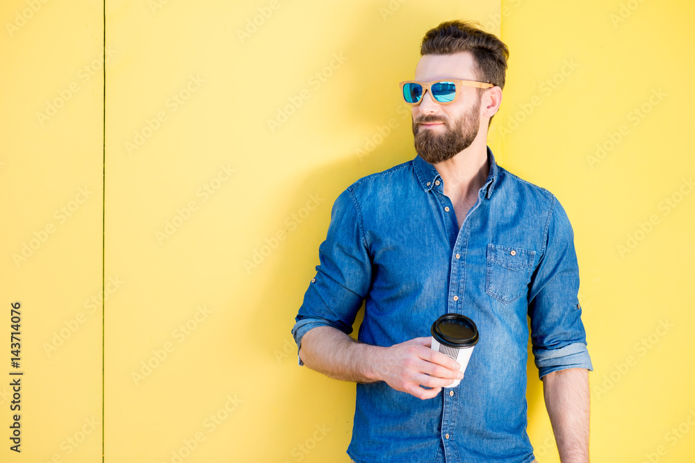 Portrait of a handsome man in blue t-shirt standing with coffee to go on the yellow background