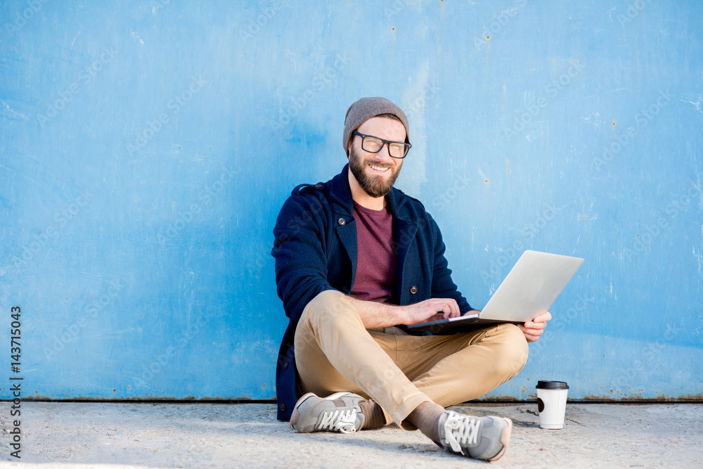 Stylish man dressed casual in sweater and hat working with laptop sitting near the blue wall backgro