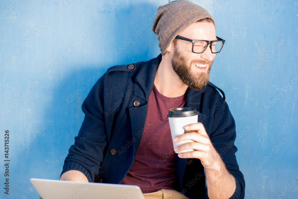 Stylish man dressed casual in sweater and hat working with laptop sitting with coffee near the blue 