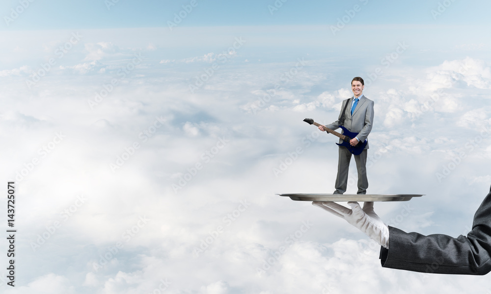 Businessman on metal tray playing electric guitar against blue sky background