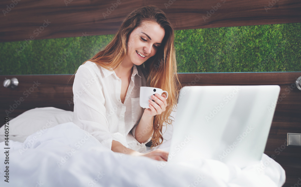 Pretty woman on bed in modern apartment using laptop and drinking coffee after wake up