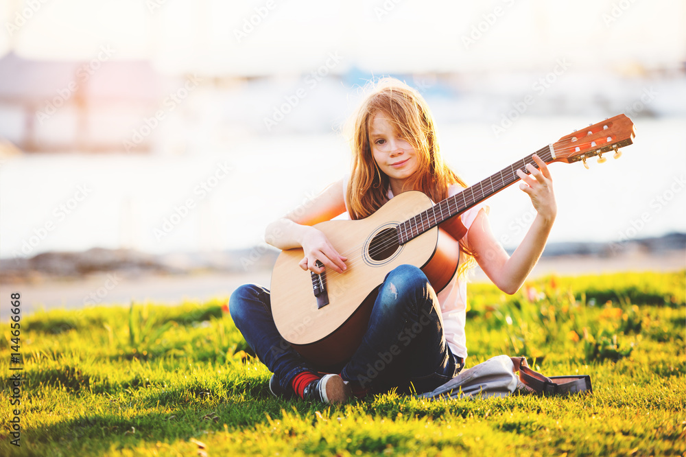 Outdoor portrait of adorable 9 year old kid girl playing guitar outdoors, sitting on bright green la