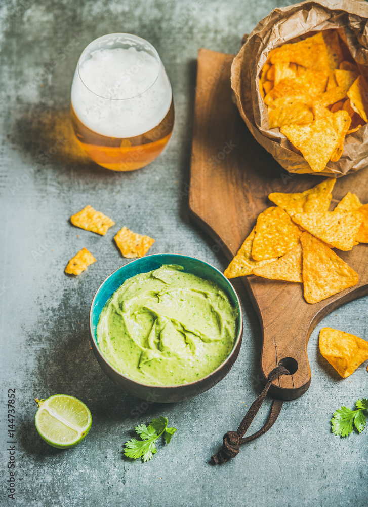 Mexican corn chips, fresh guacamole sauce and glass of beer on wooden serving board over grey concre