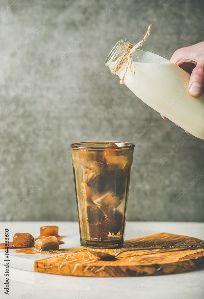 Mans hand ready to pour milk from bottle to glass with morning coffee ice cubes on olive wood and ma