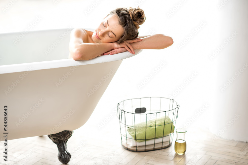 Beautiful young woman relaxing lying in the retro bathtube in the white bathroom