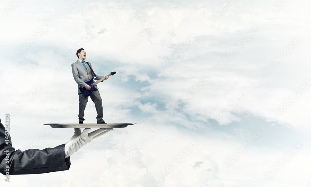 Businessman on metal tray playing electric guitar against blue sky background