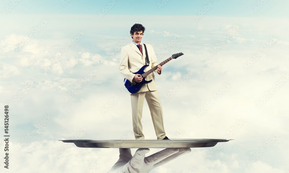 Businessman on metal tray playing electric guitar against blue sky background