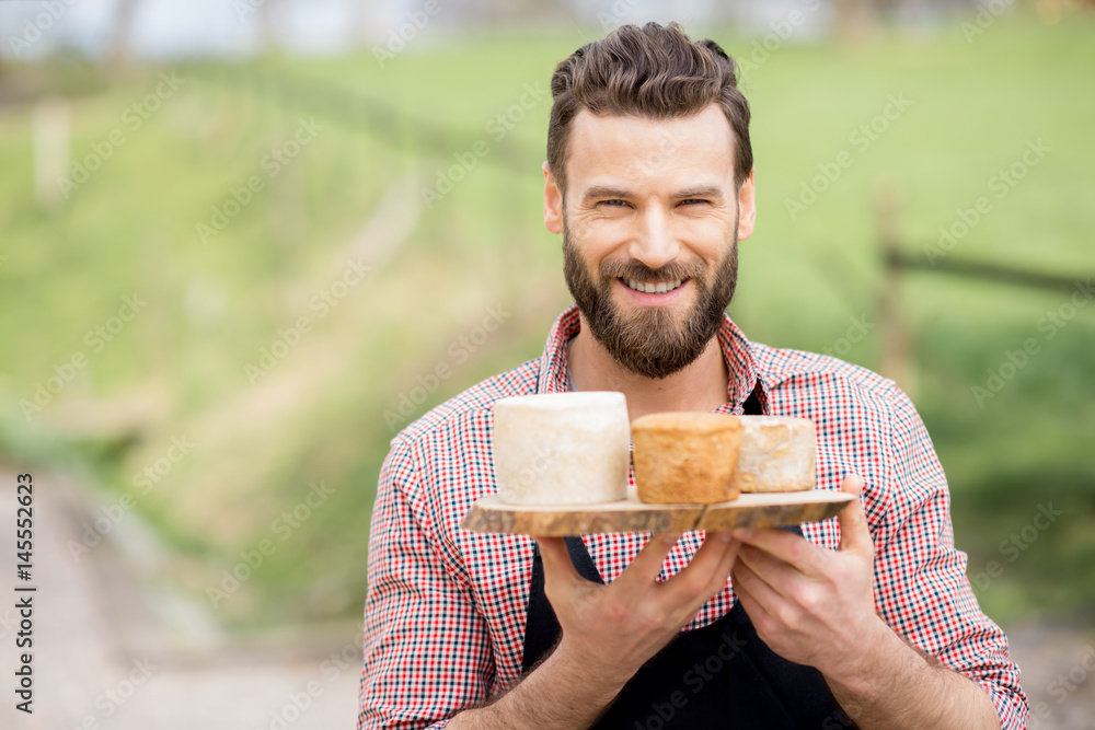 Portrait of a handsome farmer in apron standing with goat cheeses outdoors on the meadow background