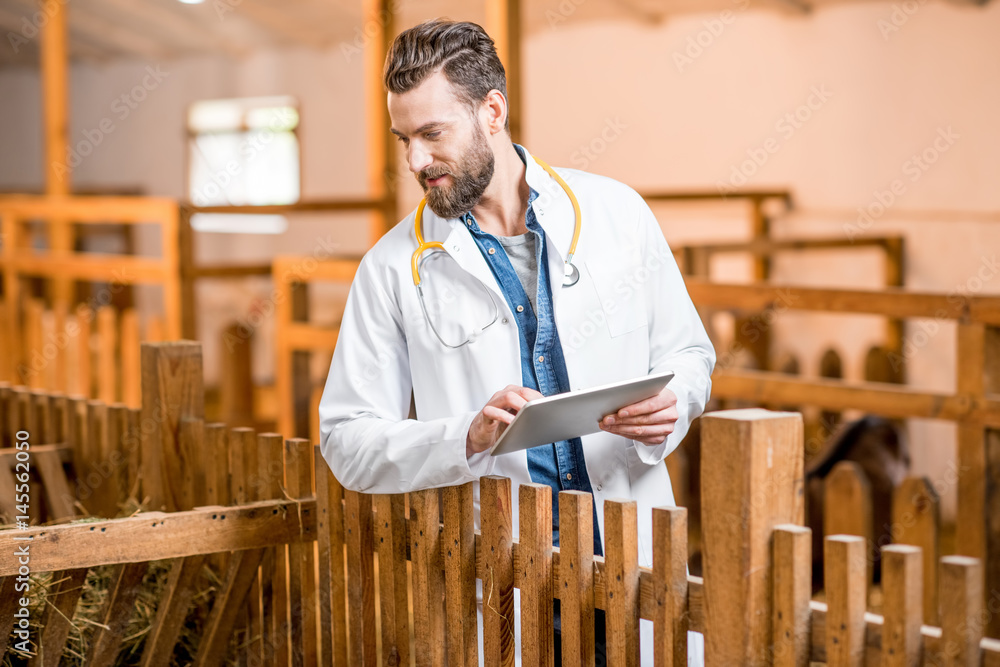 Handsome doctor in medical gown with digital tablet taking care about goats at the barn