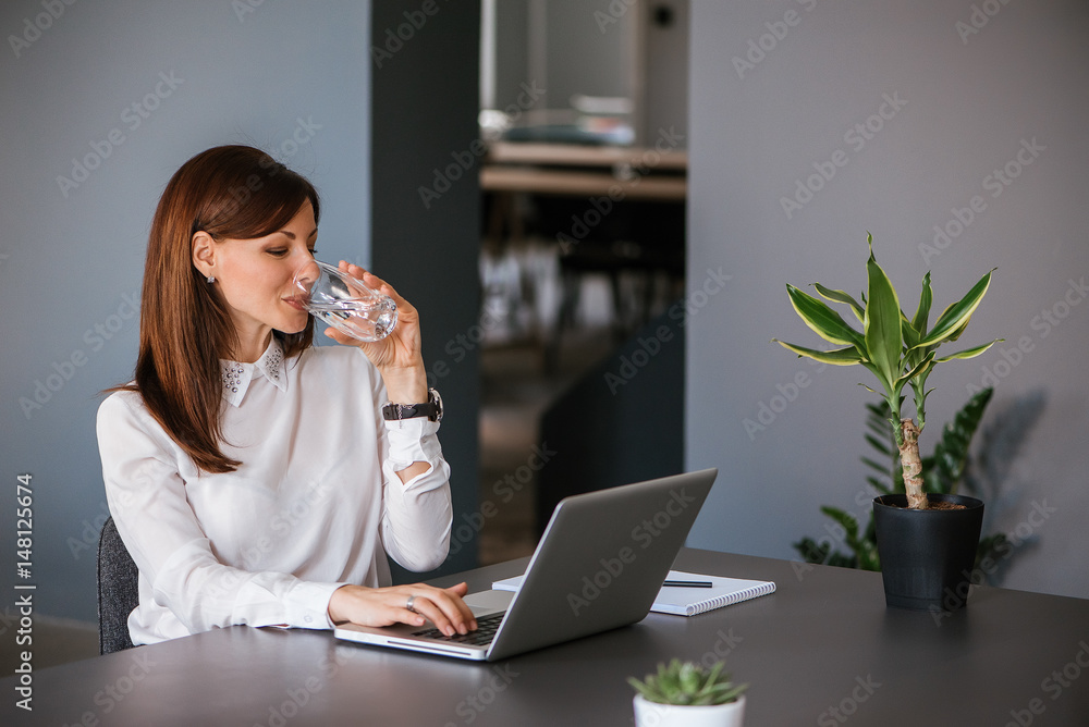 Stay hydrated. Pretty young woman in the office drinking water while working. Drinking from glass.