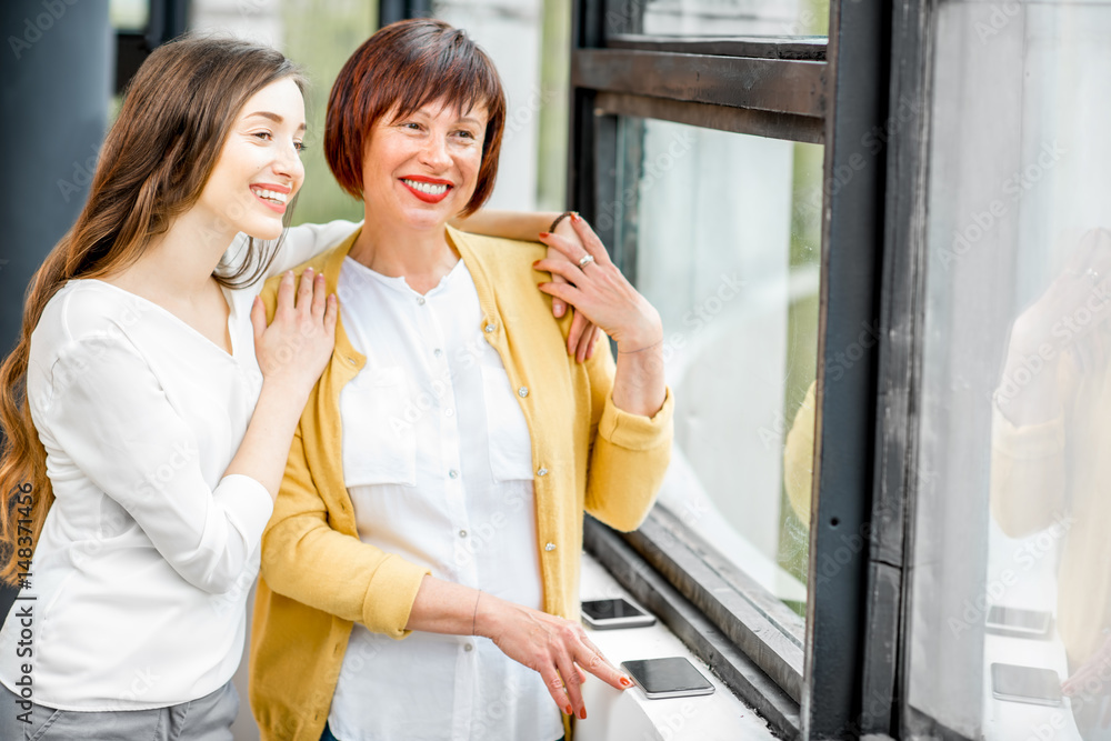 Portrait of a smiling mother and daughter standing together indoors near the window