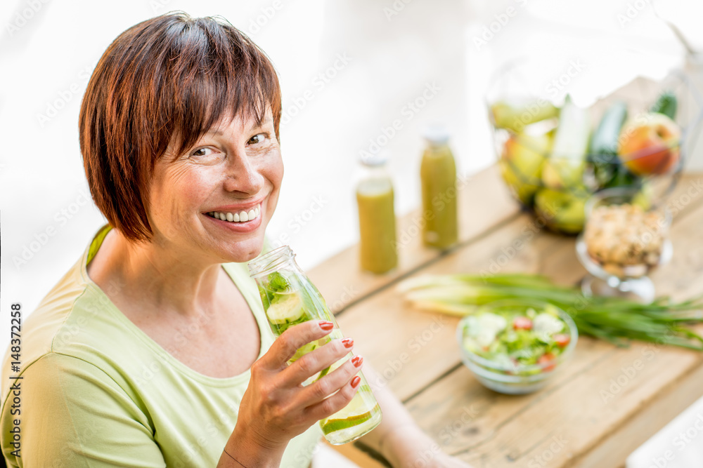 Older woman in green t-shirt drinking fresh detox drink from the bottle indoors