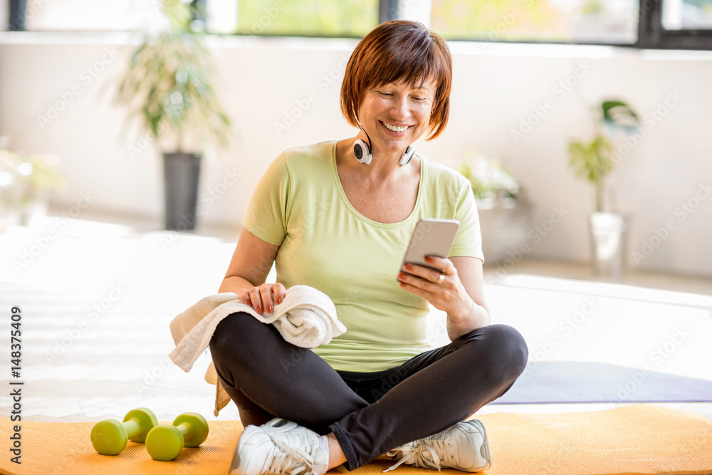 Older woman using smartphone after sports training sitting indoors at home or gym