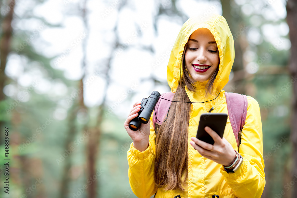 穿着黄色雨衣的年轻女子在绿色松树f中行走时使用智能手机进行定向