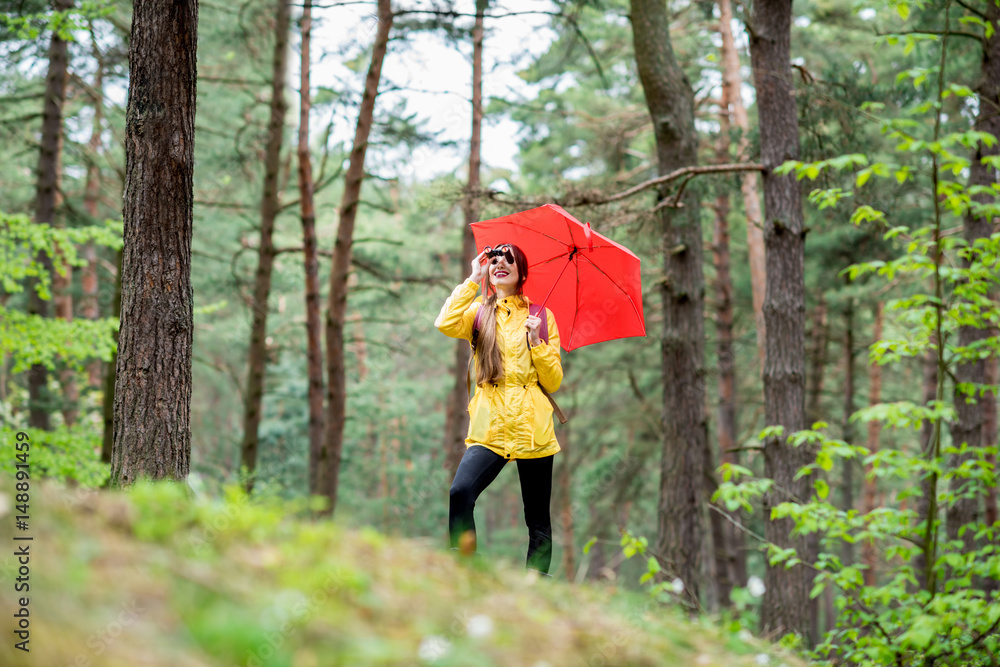 身穿黄色雨衣的年轻女子带着双筒望远镜和红色雨伞在松林中徒步旅行