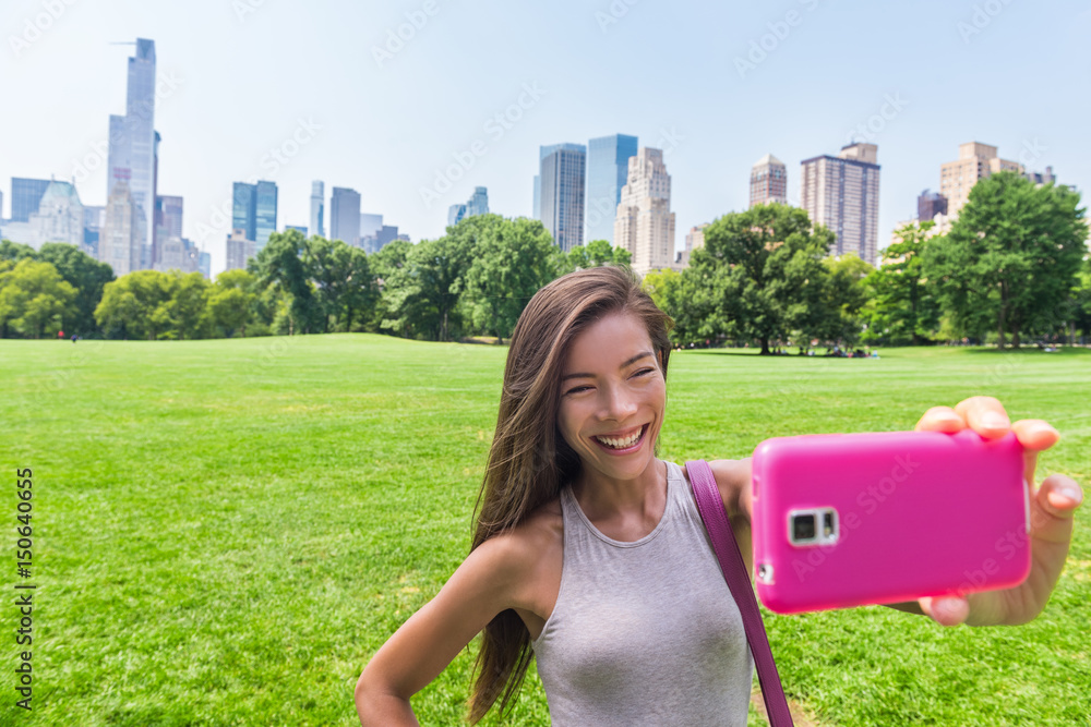 Happy Asian woman taking smartphone selfie photo with mobile phone at sheep meadow, Central park. Gi