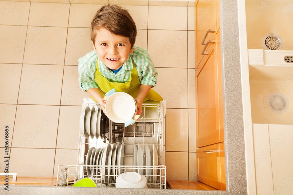 Happy kid boy taking out dishes from dishwasher