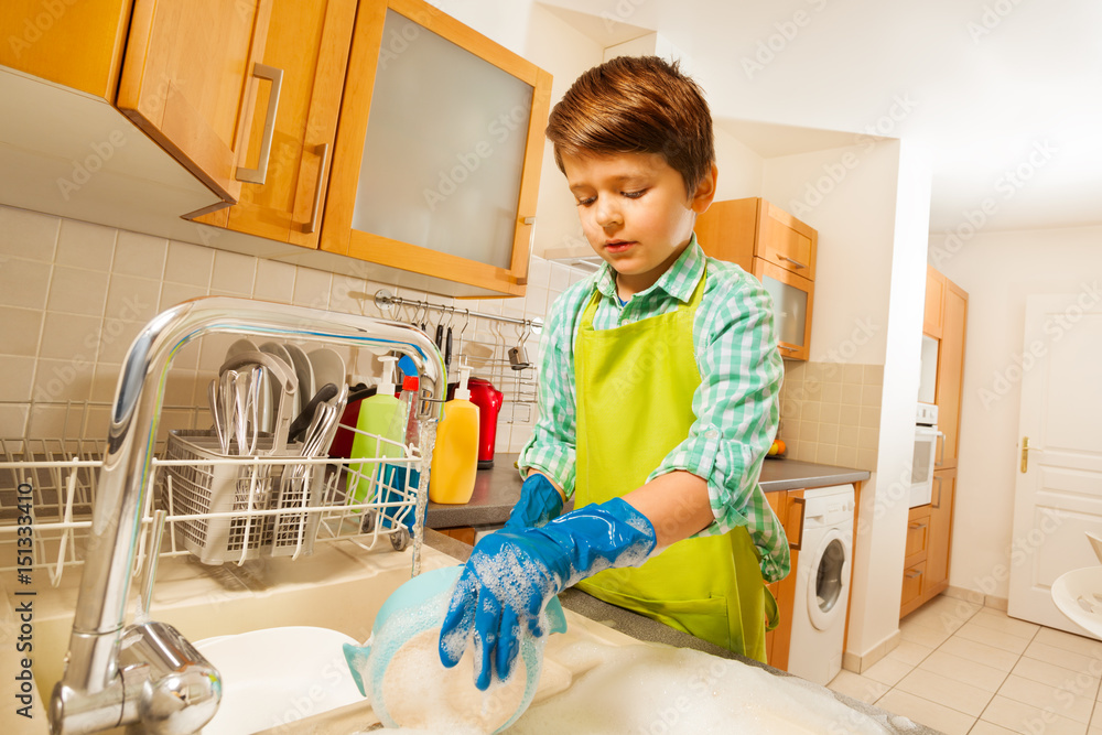 Boy doing the dishes under running water in sink