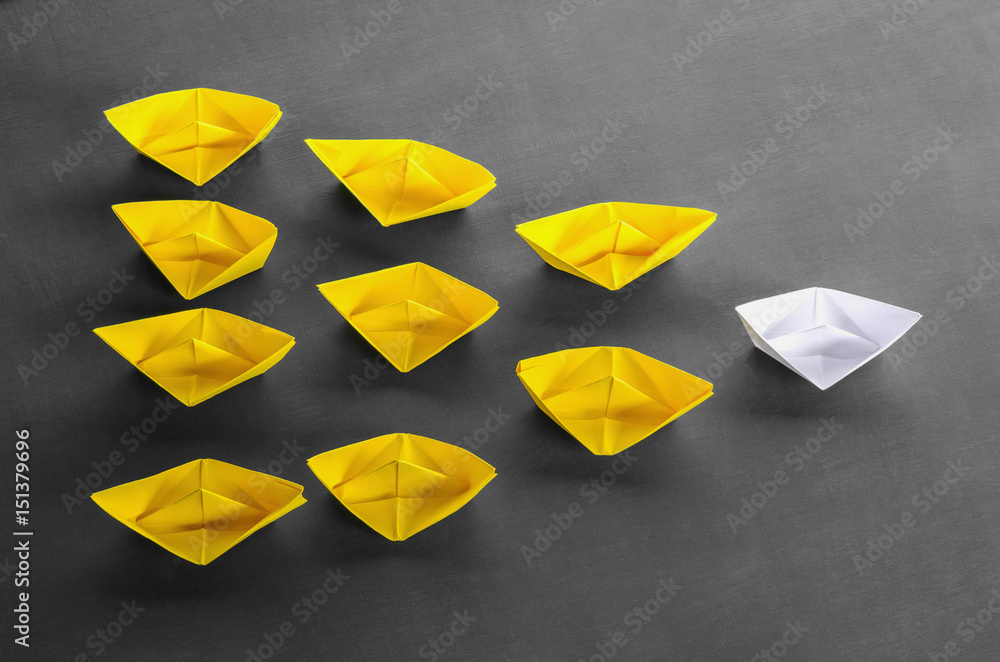 Leadership concept white leader paper boat standing out from the crowd of yellow boats on black wood