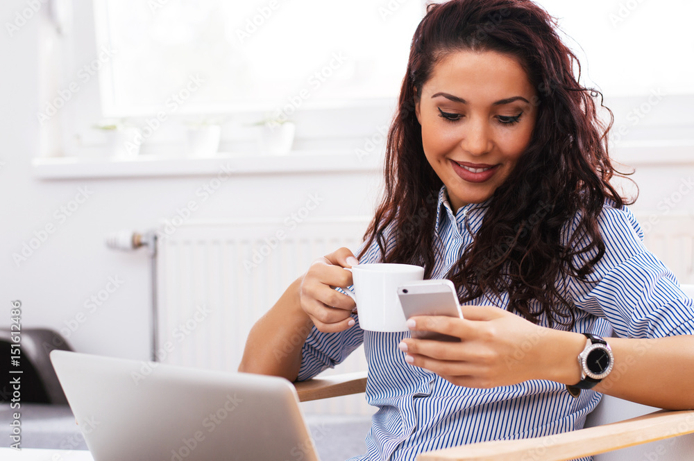 Young woman sitting next to window and texting on phone while drinking morning coffee or tea.