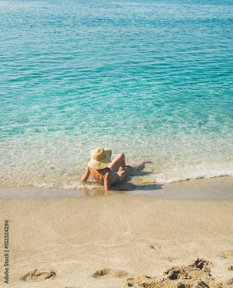 Beautiful slim senior woman tourist in bikini and hat lying on sand enjoying clear sea waters at Med