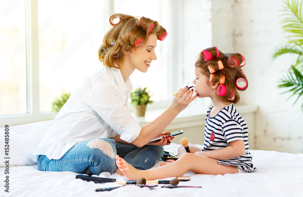 Mother and baby daughter in curlers  doing makeup on bed