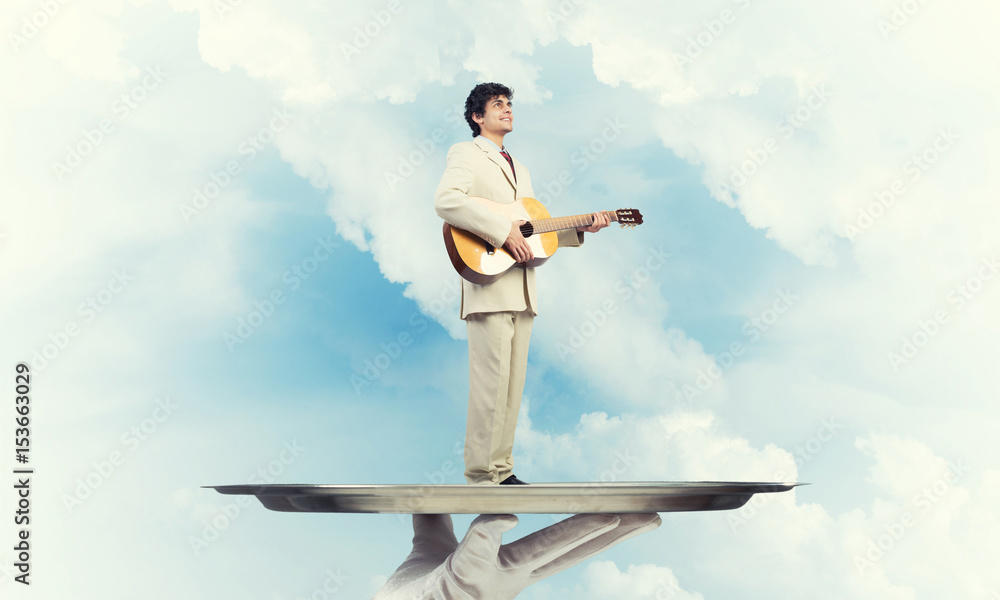 Businessman on metal tray playing acoustic guitar against blue sky background