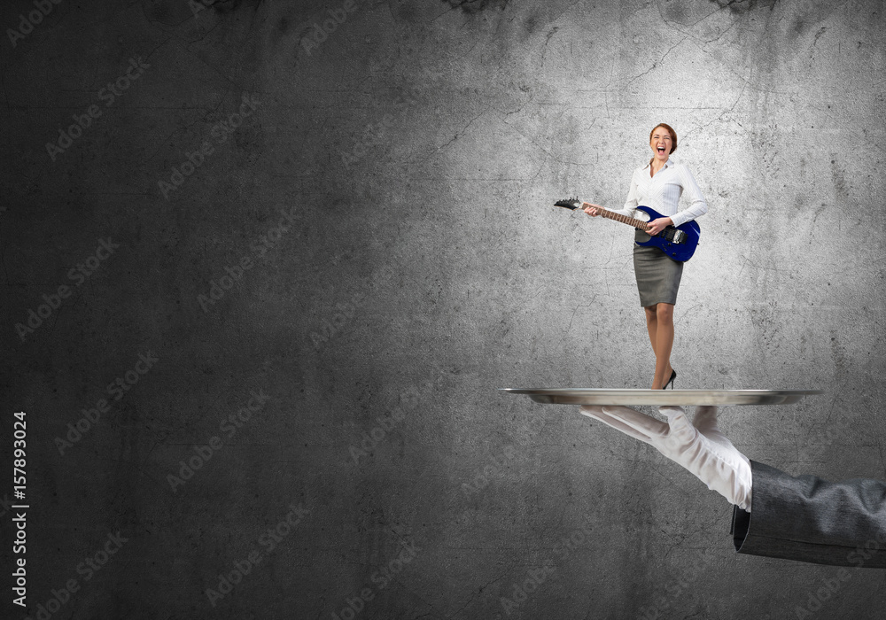 Attractive businesswoman on metal tray playing electric guitar against concrete wall background