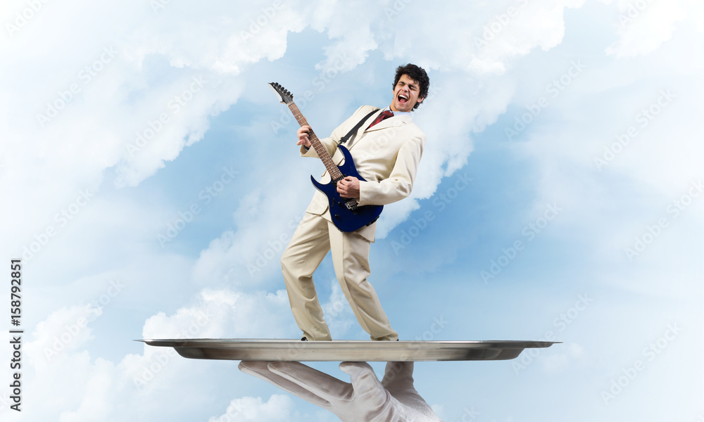 Businessman on metal tray playing electric guitar against blue sky background