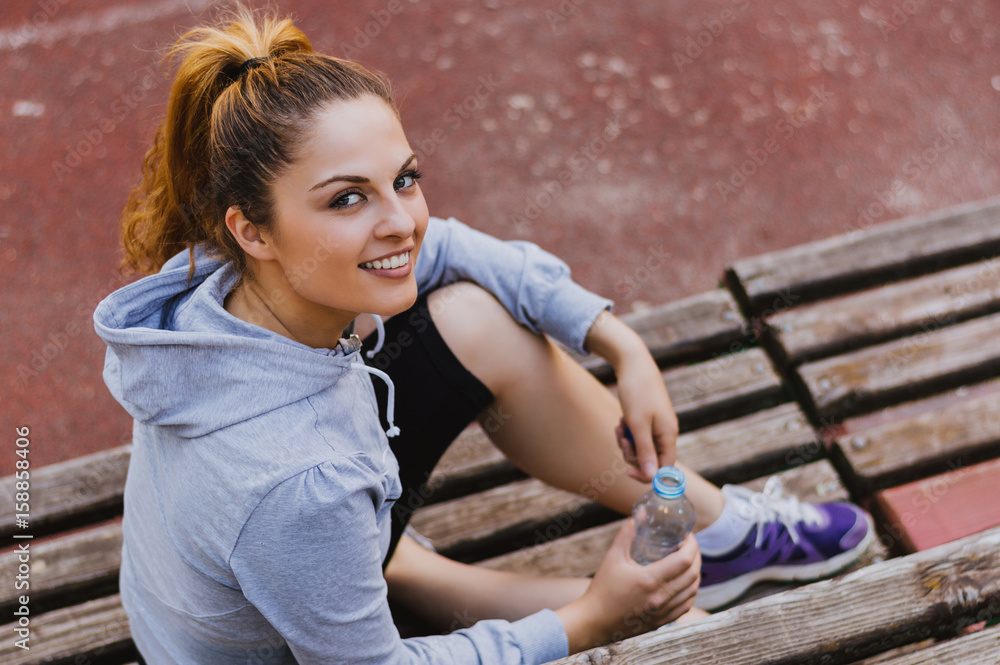 Sporty and attractive young woman sitting down on a wooden bench in the city, taking a break from ex