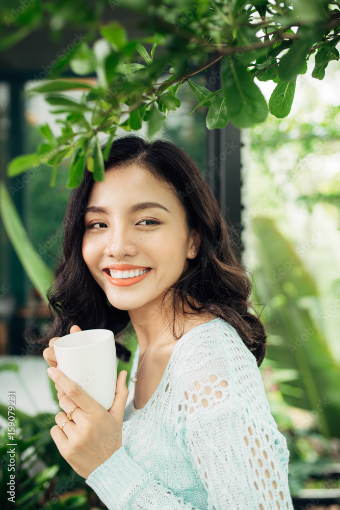 快乐的年轻亚洲女人，手里拿着马克杯，站在阳台上喝咖啡。