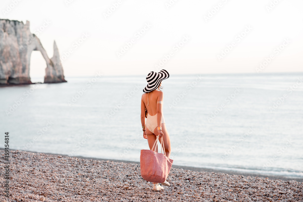 Woman in hat and swimsuit standing back with beach bag enjoying great view on the ocean during the s