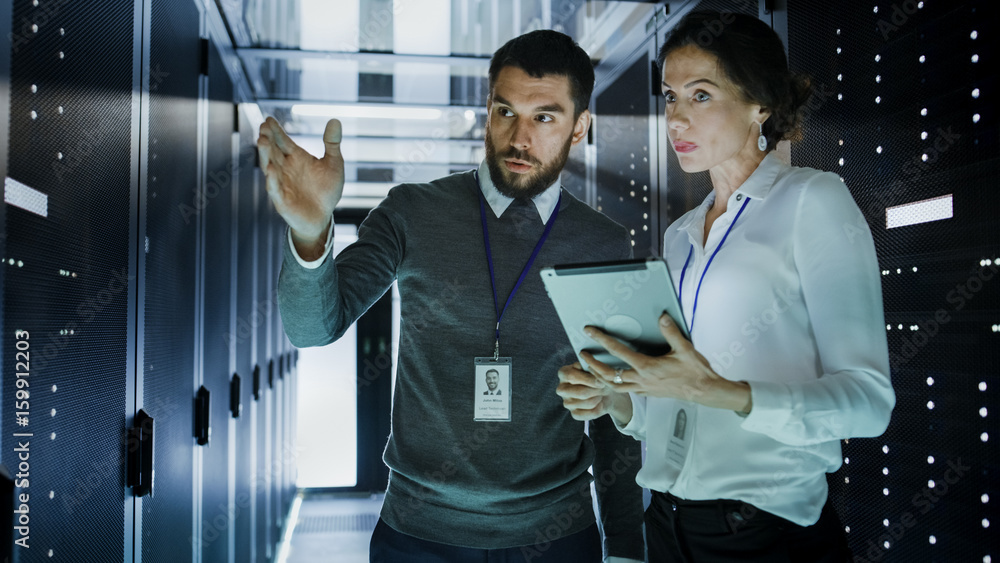 Two IT Engineers in Data Center / Server Room, Talking, one of them Pointing Forward, Other Holds Ta