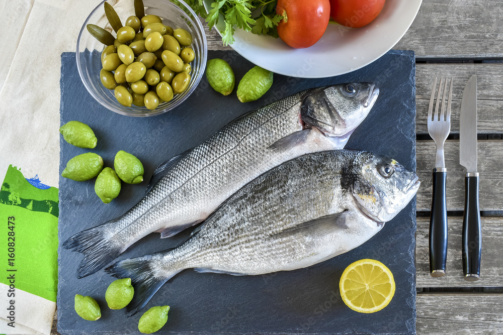 Two ready to cook raw fish with lemon and olives on stone slate board.