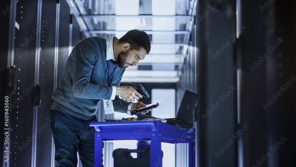 IT Engineer with Tool Cart Working on a Laptop Computer, he Holds a Hard Drive. He Stands at a Corri