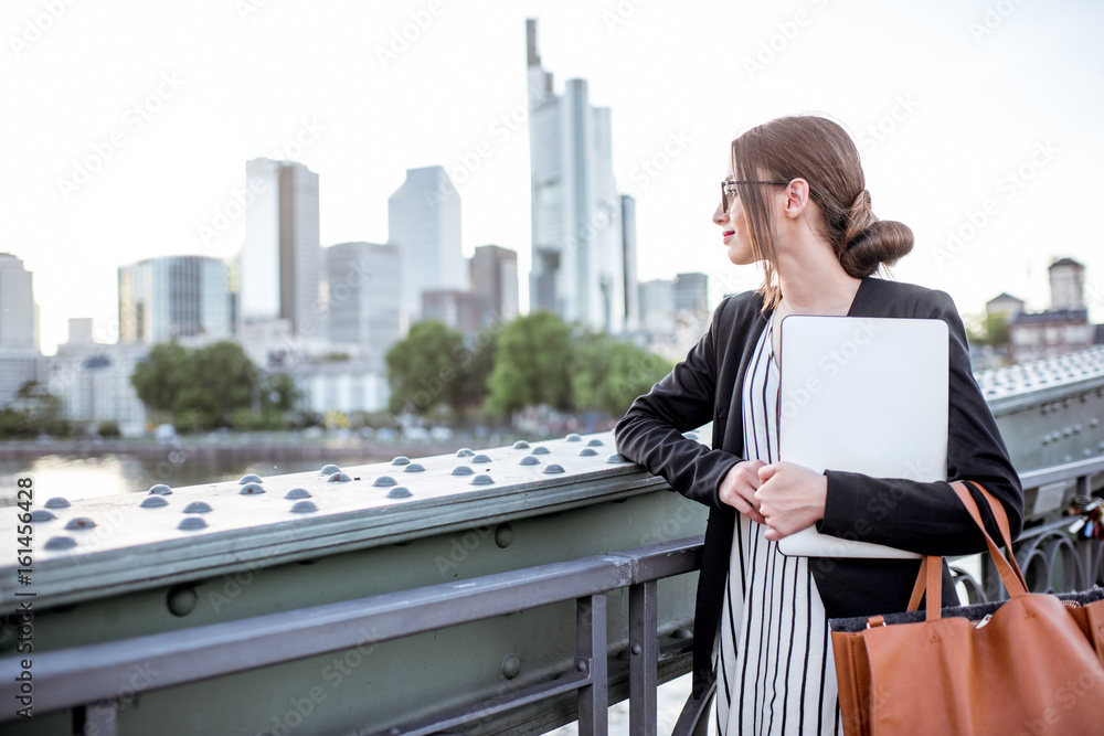 Portrait of a young businesswoman with laptop standing on the bridge in Frankfurt city